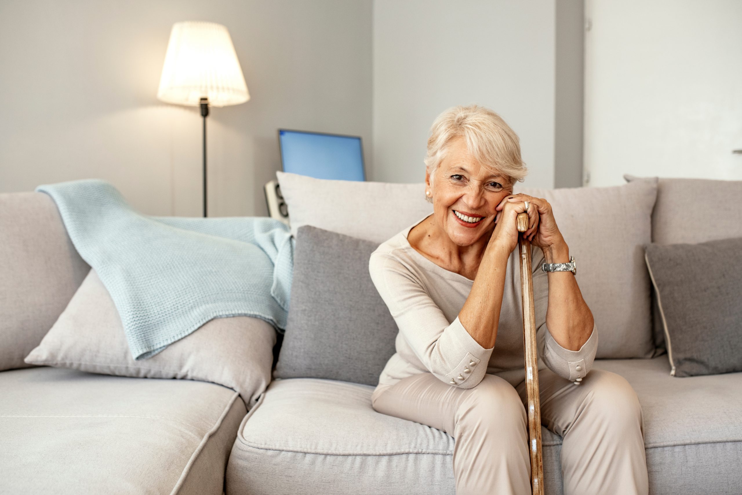 Photo of Retired short hair old woman with her walking stick at home looking at camera during the day while sitting on sofa. Senior gray hair women supporting on a walking cane at home.