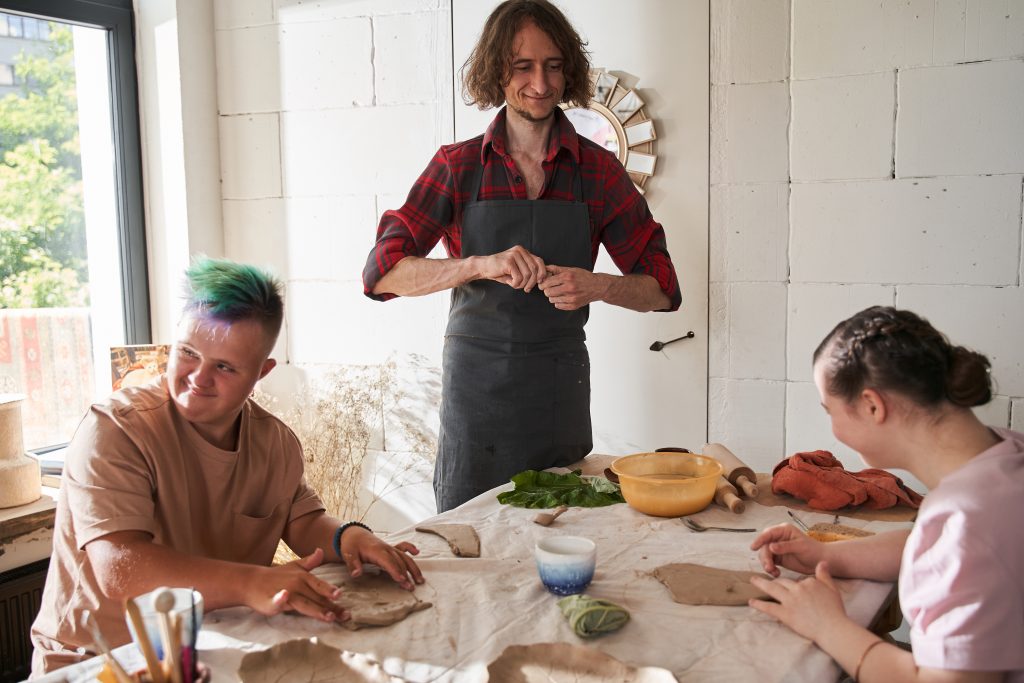 Working process. Full length view of the happy sculptor wearing apron smiling while teaching his master class for the people with special needs. Stock photo