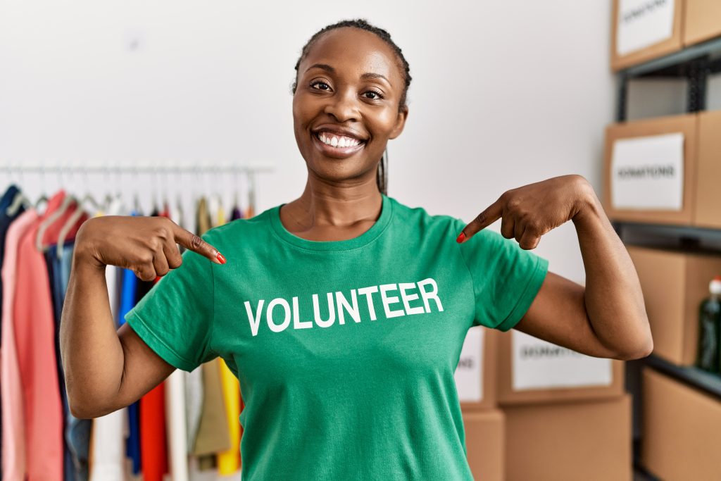 Young african american woman smiling happy pointing with finger to volunteer t shirt at charity center.
