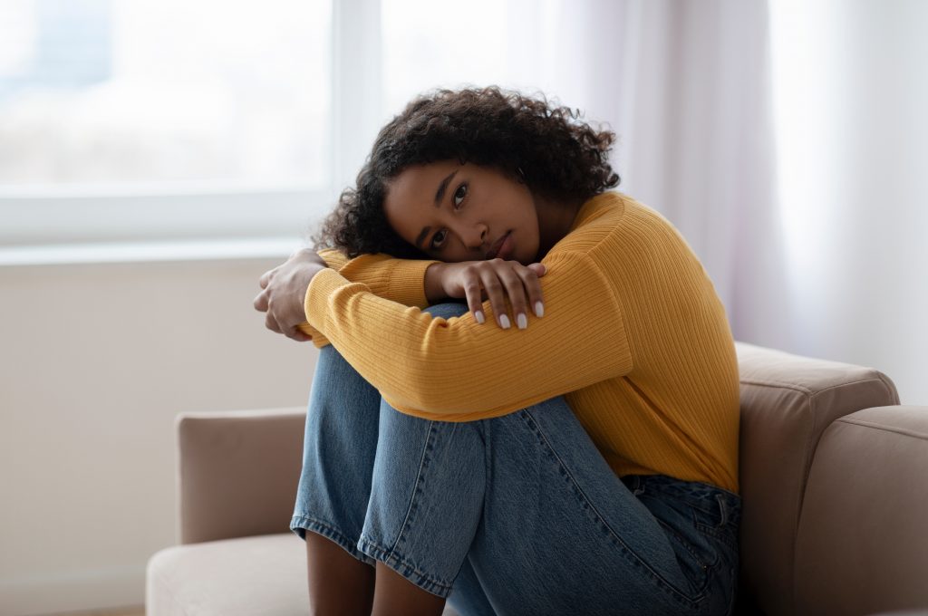 Young depressed black woman hugging her knees on sofa at home