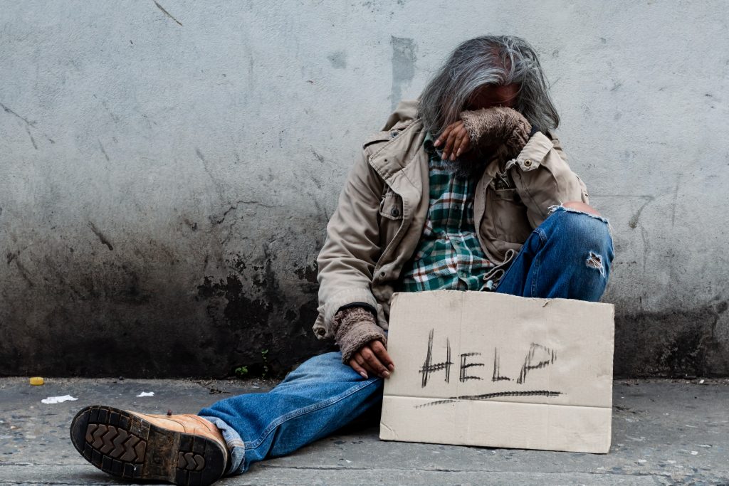 Homeless, long-haired Asian man sits hopelessly leaning against a wall as there is no one to help him with work and food in his hand holding a sign for help.